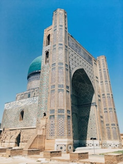 A grand historical building with intricate mosaic patterns and geometric designs covers the facade. The structure features a large archway and a prominent turquoise dome, characteristic of Islamic architecture. The brickwork and decorative tiles exhibit a blend of earthy and vibrant colors, reflecting meticulous craftsmanship.