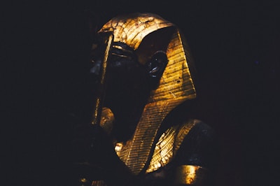 A statue of a pharaoh is partially illuminated in dramatic lighting, highlighting the gold and dark material of the figure. The pharaoh is adorned with a headdress and holds a staff.
