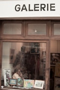 Inside an art gallery, a person is seated and surrounded by various framed artworks. Some paintings and prints are displayed on easels and the walls. The scene is viewed through a window bearing the word 'GALERIE' above it.