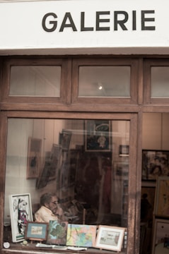 Inside an art gallery, a person is seated and surrounded by various framed artworks. Some paintings and prints are displayed on easels and the walls. The scene is viewed through a window bearing the word 'GALERIE' above it.
