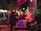 A lively nighttime street scene with people gathered outside a café. The atmosphere is vibrant with glowing neon lights reflecting on the street, creating a colorful and energetic vibe. A group of people are sitting and standing, some engaged in conversation, while others enjoy hookah, visible through the smoke drifting upwards.