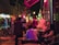 A lively nighttime street scene with people gathered outside a café. The atmosphere is vibrant with glowing neon lights reflecting on the street, creating a colorful and energetic vibe. A group of people are sitting and standing, some engaged in conversation, while others enjoy hookah, visible through the smoke drifting upwards.