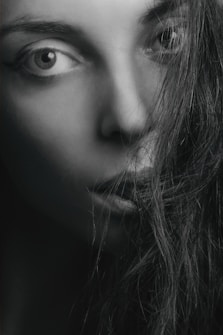 A close-up black and white portrait of a person with intense eyes staring directly. The face is partially obscured by strands of long, dark hair, creating a dramatic and mysterious effect. The lighting highlights the contours of the face and features.