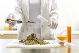 A person wearing a lab coat and gloves is handling dried plant material on a white cutting board. They are using a spatula to place some of it into a test tube or small vial. On the table is a graduated cylinder filled with an orange liquid and a round-bottom flask partially filled with a similar substance.