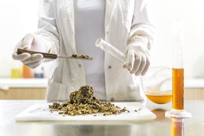 A person wearing a lab coat and gloves is handling dried plant material on a white cutting board. They are using a spatula to place some of it into a test tube or small vial. On the table is a graduated cylinder filled with an orange liquid and a round-bottom flask partially filled with a similar substance.