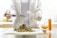 A person wearing a lab coat and gloves is handling dried plant material on a white cutting board. They are using a spatula to place some of it into a test tube or small vial. On the table is a graduated cylinder filled with an orange liquid and a round-bottom flask partially filled with a similar substance.