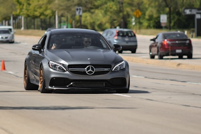 A sleek, dark-colored luxury car is driving on a multi-lane road, with a driver visible through the windshield. Several other vehicles can be seen in the background, and the surrounding area is lined with lush green trees.