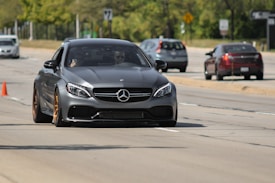 A sleek, dark-colored luxury car is driving on a multi-lane road, with a driver visible through the windshield. Several other vehicles can be seen in the background, and the surrounding area is lined with lush green trees.