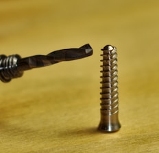 A close-up of a drill bit positioned near a metal screw, placed on a blurred gold surface. The drill bit features a spiral fluted design, while the screw displays a shiny, threaded surface with a rounded head.