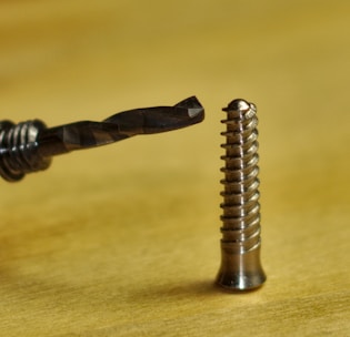 A close-up of a drill bit positioned near a metal screw, placed on a blurred gold surface. The drill bit features a spiral fluted design, while the screw displays a shiny, threaded surface with a rounded head.