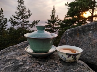 A green tea set is placed on a large rock with a scenic backdrop of evergreen trees and a setting sun. A lidded tea cup sits next to a small bowl containing tea, both featuring a traditional design. The atmosphere is calm and natural.