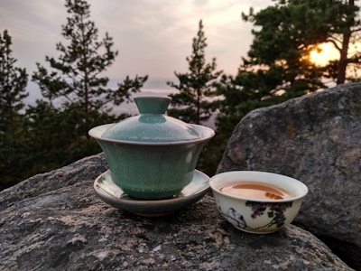 A green tea set is placed on a large rock with a scenic backdrop of evergreen trees and a setting sun. A lidded tea cup sits next to a small bowl containing tea, both featuring a traditional design. The atmosphere is calm and natural.