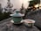 A green tea set is placed on a large rock with a scenic backdrop of evergreen trees and a setting sun. A lidded tea cup sits next to a small bowl containing tea, both featuring a traditional design. The atmosphere is calm and natural.