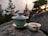 A green tea set is placed on a large rock with a scenic backdrop of evergreen trees and a setting sun. A lidded tea cup sits next to a small bowl containing tea, both featuring a traditional design. The atmosphere is calm and natural.