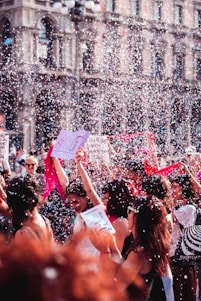 A vibrant street protest or celebration features a group of people gathered together, surrounded by confetti and holding signs with messages. One prominent sign has the hashtag #LOVE. The crowd is lively, marked by bright colors and energy, suggesting a festive atmosphere. The background shows an ornate building with arches and windows.