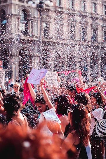 A vibrant street protest or celebration features a group of people gathered together, surrounded by confetti and holding signs with messages. One prominent sign has the hashtag #LOVE. The crowd is lively, marked by bright colors and energy, suggesting a festive atmosphere. The background shows an ornate building with arches and windows.