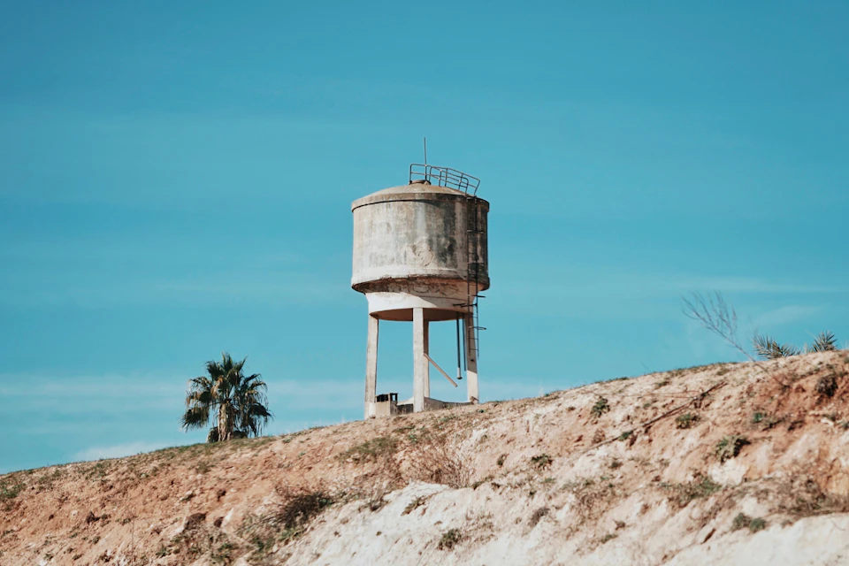 The image depicts a serene rural landscape with a large, gray water tower standing on a hillside. The water tower is surrounded by a dirt field and a palm tree, adding to the tranquil atmosphere. In the distance, there are more trees visible against the blue sky, creating depth in the scene.