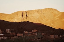 A rugged mountain landscape bathed in golden light during what appears to be either sunrise or sunset. Shadows are cast on parts of the mountain, creating a stark contrast between light and dark areas. Below the mountain, a small village with flat-roofed buildings sits quietly at the base, constructed in earthy tones to blend with the natural surroundings.