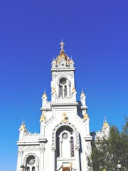 A grand, ornate church with a prominent gold and white facade. The architecture includes intricate details, arches, and a large gold dome with a cross on top. The building stands against a clear blue sky, with some greenery visible at the side.