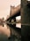 A large, intricately designed bridge arches over a reflective body of water, creating a symmetrical image with its reflection. The structure is composed of metal and stone elements, showcasing detailed engineering work. In the background, there is a cityscape with various tall buildings under a cloudy sky, giving an urban feel.