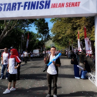 A man is standing under a start-finish banner holding inflatable sticks, with a crowd in the background. The event appears to be a healthy walk or road event, evidenced by people wearing casual sportswear and red and white attire. Vehicles are seen parked, including an ambulance, adding a sense of safety and organization. Trees line the road, providing a natural backdrop.