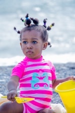 A young child sits at the beach wearing a bright pink and white striped swimsuit adorned with two turquoise seahorses. The hairstyle features several small puffs, each tied with colorful bands. The child is holding a yellow shovel and appears to be playing with a matching yellow bucket.