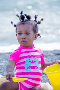 A young child sits at the beach wearing a bright pink and white striped swimsuit adorned with two turquoise seahorses. The hairstyle features several small puffs, each tied with colorful bands. The child is holding a yellow shovel and appears to be playing with a matching yellow bucket.