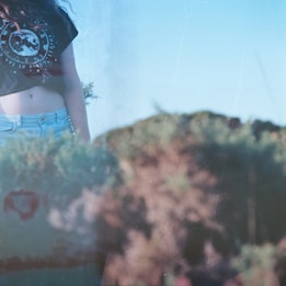 A person wearing a black crop top with a celestial design and light blue denim jeans stands partially in view. The background is a blurred outdoor scene featuring greenery and possibly shrubs or bushes under a clear blue sky.