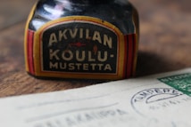 An old ink bottle with a label that reads 'AKVILAN KOULU-MUSTETTA' is placed on a wooden surface next to a piece of paper with stamps and postmarks, including the word 'TAMPEKE'. The ink bottle has a vintage design with red and yellow stripes around the label.