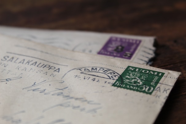 Two vintage envelopes are stacked on a wooden surface, each with distinctive postmarks and colorful stamps. The top envelope features a green stamp with the word 'SUOMI' and 'FINLAND,' while the envelope below has a purple stamp.