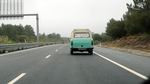 A vintage turquoise van drives down a deserted highway surrounded by greenery on a cloudy day. The van's windows are decorated with various colorful stickers. The scene conveys a sense of travel and adventure.