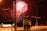 Fireworks illuminate the night sky over a street with orange traffic barrels and street lights. Two figures stand near a utility box, blurred from movement. A car's lights streak by, creating a dynamic urban scene.