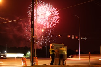 Fireworks illuminate the night sky over a street with orange traffic barrels and street lights. Two figures stand near a utility box, blurred from movement. A car's lights streak by, creating a dynamic urban scene.