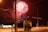 Fireworks illuminate the night sky over a street with orange traffic barrels and street lights. Two figures stand near a utility box, blurred from movement. A car's lights streak by, creating a dynamic urban scene.