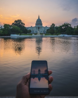 A hand holding a smartphone captures the reflection of the Capitol building in a body of water at sunset. The sky displays a gradient of warm hues, and lush trees frame the structure.