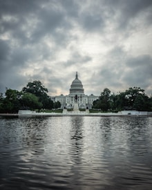 A large, neoclassical government building with a dome is framed by lush green trees. The scene is reflected in a calm body of water in the foreground, under an overcast sky.