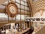 An ornate, large clock is prominently displayed, featuring Roman numerals and intricate metalwork. The setting is a grand, historic interior with high ceilings, detailed architectural elements, and large arched windows. Below, people are walking and interacting in a museum-like environment, with information desks and exhibitions visible.