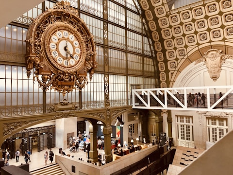 An ornate, large clock is prominently displayed, featuring Roman numerals and intricate metalwork. The setting is a grand, historic interior with high ceilings, detailed architectural elements, and large arched windows. Below, people are walking and interacting in a museum-like environment, with information desks and exhibitions visible.