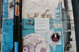 A textured urban wall with layers of torn posters and graffiti, featuring a prominent black pole with colorful stickers, a white paper with text, and a sticker advertising street art tours. The wall displays varied patterns and remnants of old advertisements, creating an eclectic and somewhat chaotic visual.