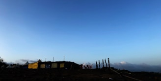 A rural landscape featuring a row of tents or makeshift structures with some yellow tarps. Wooden stakes and plastic chairs are scattered around, set against a backdrop of a wide, open sky transitioning from blue to light.