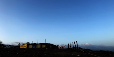 A rural landscape featuring a row of tents or makeshift structures with some yellow tarps. Wooden stakes and plastic chairs are scattered around, set against a backdrop of a wide, open sky transitioning from blue to light.