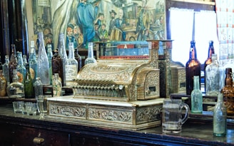A vintage, ornate cash register surrounded by a variety of old glass bottles, set against a backdrop of an antique-looking mural. The cash register features intricate designs and metallic embellishments, sitting on a dark wooden surface alongside empty glassware.