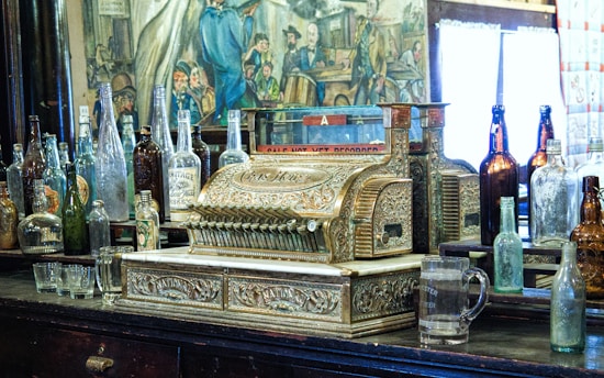 A vintage, ornate cash register surrounded by a variety of old glass bottles, set against a backdrop of an antique-looking mural. The cash register features intricate designs and metallic embellishments, sitting on a dark wooden surface alongside empty glassware.
