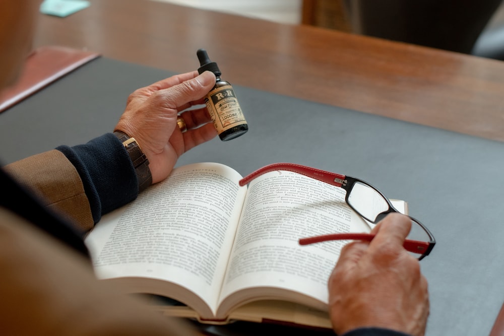 person holding small drop bottle and eyeglasses while reading book