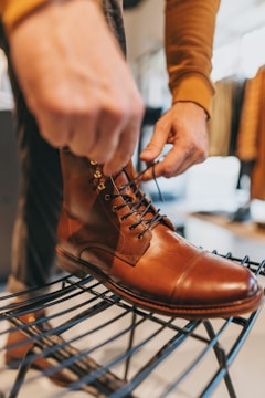 A hand is tying the laces of a polished brown leather boot placed on a black metal rack in a well-lit room. The boot has a classic design with a smooth, shiny texture and detailed stitching. The background shows blurred clothing items, suggesting a shopping or retail environment.