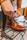 A hand is tying the laces of a polished brown leather boot placed on a black metal rack in a well-lit room. The boot has a classic design with a smooth, shiny texture and detailed stitching. The background shows blurred clothing items, suggesting a shopping or retail environment.