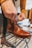 A hand is tying the laces of a polished brown leather boot placed on a black metal rack in a well-lit room. The boot has a classic design with a smooth, shiny texture and detailed stitching. The background shows blurred clothing items, suggesting a shopping or retail environment.