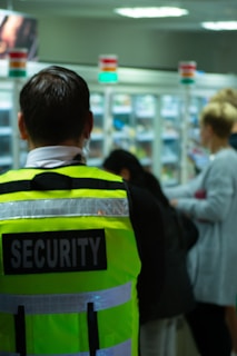 A person wearing a high-visibility security vest stands in a retail environment. The background is slightly blurred, showing other people and shelves stocked with products.