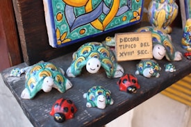 A collection of colorful ceramic figures arranged on a shelf. The figures include turtles with intricate patterns in shades of green, yellow, and blue, and red ladybugs with black spots. A small wooden sign reads 'DECORO TIPICO DEL XVIII SEC.' The background includes a decorative ceramic piece with vibrant hues and abstract designs.