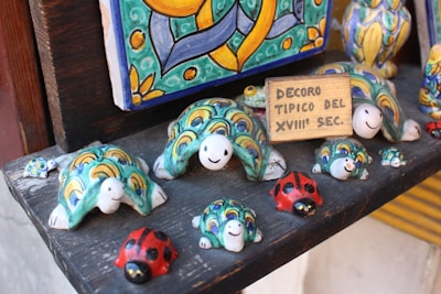A collection of colorful ceramic figures arranged on a shelf. The figures include turtles with intricate patterns in shades of green, yellow, and blue, and red ladybugs with black spots. A small wooden sign reads 'DECORO TIPICO DEL XVIII SEC.' The background includes a decorative ceramic piece with vibrant hues and abstract designs.